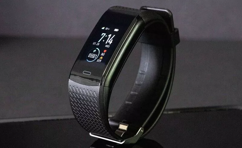 This image shows a black SmartTrack placed on a reflective surface. The screen displays the time '7:14', the date Wednesday, July 29, the step count (05501), the heart rate (043), as well as other health and fitness-related information. The strap has a textured design, and a touch button is visible beneath the screen.