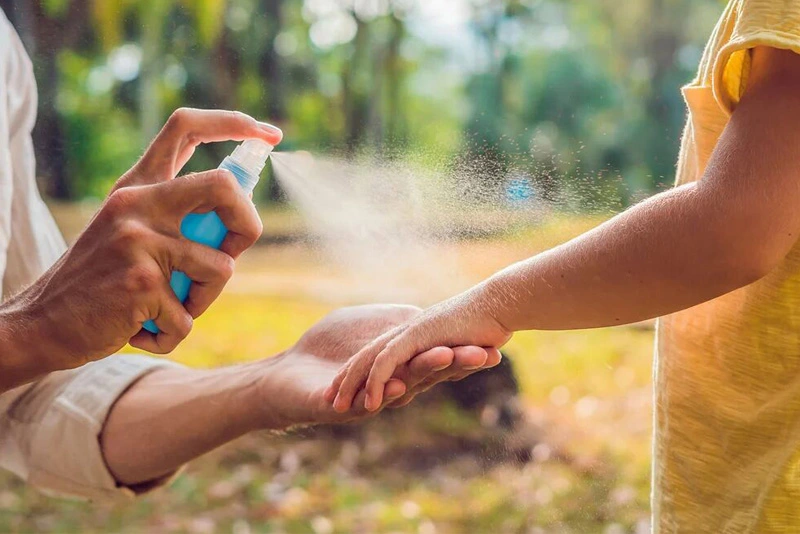 L'image montre une personne vaporisant un produit anti-moustique sur le bras d'un enfant, en plein air, dans un cadre naturel. Le spray est tenu dans une main et appliqué soigneusement sur l'avant-bras de l'enfant, qui porte un t-shirt jaune. L'arrière-plan est flou, suggérant une scène en extérieur, probablement dans un parc ou un jardin.
